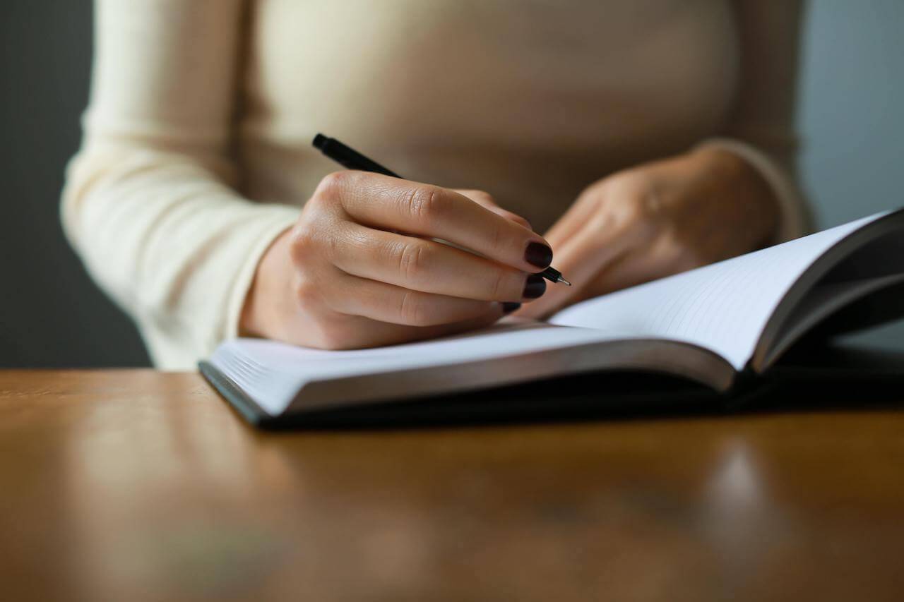 Image of a woman's hand holding a pen writing in a journal. For article by Stephanie Sarkis on Psychology Today "Consider Skipping New Year's Resolutions in 2021" PC: ava-sol-JaUn2B6smQs-unsplash